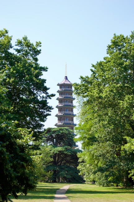 A tall, multi-tiered pagoda situated behind dense green trees in a park setting, with a narrow paved pathway leading towards it. The pagoda has several distinct levels with traditional curved roofs, a
