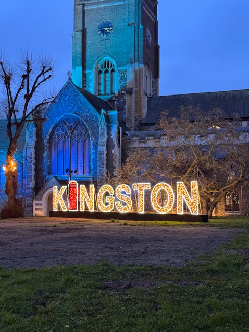 A large illuminated sign spelling 'KINGSTON' in bright, colourful letters is positioned on the grass in front of a historic stone church with tall stained-glass windows, which is lit with blue lightin