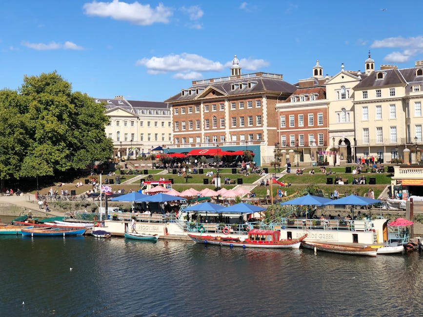 A scenic riverside scene in Kingston upon Thames featuring a row of small boats docked along the water's edge, some covered with blue and pink umbrellas, with a large tree on the left providing shade. Behind the boats, there is a grassy area with people sitting on benches and ledges, enjoying the outdoor setting. In the background, multi-storey historic buildings with a mix of cream and brick facades line the riverbank, some with decorative towers and gabled roofs, creating a characteristic townscape near Kingston Station. The sky is partly cloudy with patches of blue, suggesting a pleasant day suitable for a home relocation involving packing and moving activities. The presence of boats, outdoor seating, and the established urban environment demonstrate the vibrant riverside atmosphere in Kingston, which [COMPANY_NAME] might support during house removals, offering transport and logistics services in the Kingston upon Thames area. The overall scene is calm and active, illustrating a typical day in a popular town location, with the focus on the picturesque setting supporting residential moving and furniture transport services. 