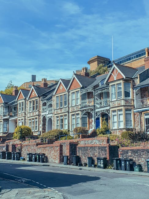 A row of Victorian-style terraced houses in Kingston upon Thames, captured during daytime with clear blue sky and scattered clouds. The houses feature decorative ironwork balconies, large sash windows, and brick facades with ornate detailing and gabled roofs. The buildings are elevated above the pavement, with brick retaining walls and small landscaped front gardens. Along the street, black wheelie bins are lined up in front of the houses, indicating typical residential waste disposal. The road is quiet, with no visible traffic or pedestrians, suggesting a tranquil neighbourhood suited for home relocation activities. This image reflects an area where professional removals services, such as those provided by Kingston upon Thames-based specialists, are often required for moving furniture and boxes during house moves near Kingston Station.