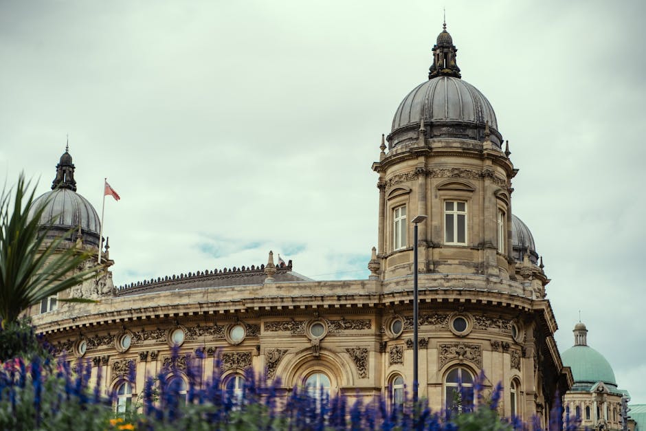 A large, historic building with a classical architectural style featuring a central statue of a seated figure holding a flag, flanked by two domed towers on either side. The structure is made of light
